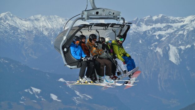 Skifahrer in einem Sessellift vor verschneiten Bergen.
