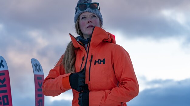 Woman in orange ski jacket with hat and skis | © Helly Hansen / Cam McLeod