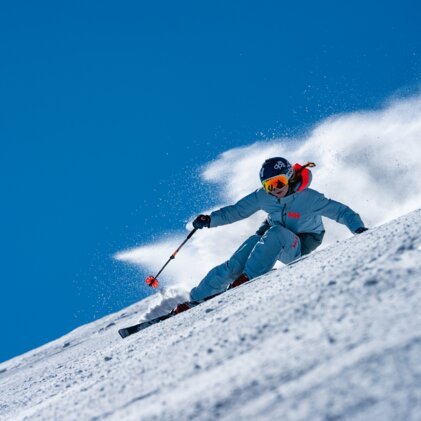 Skier carving through powder snow against a blue sky. | © Helly Hansen / Cam McLeod