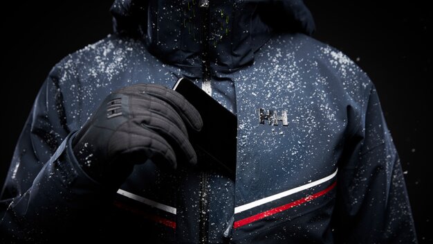 Person wearing a blue winter jacket with a black glove and putting a smartphone into the pocket. | © Helly Hansen