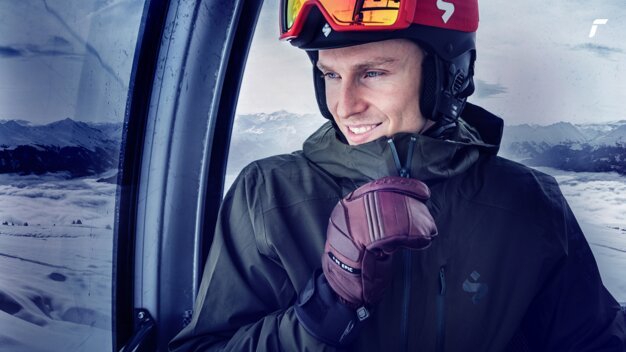 Mann im Skilift mit Helm und Handschuhen lächelt. | © Reusch