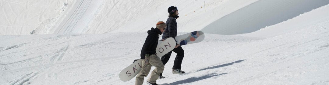 Two snowboarders carrying their boards on a snowy slope. | © SALOMON