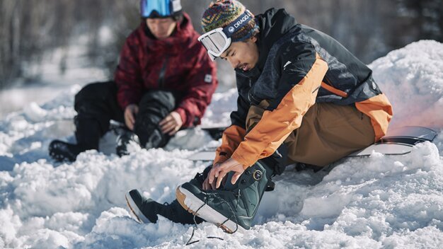 Two snowboarders putting on boots in the snow. | © SALOMON