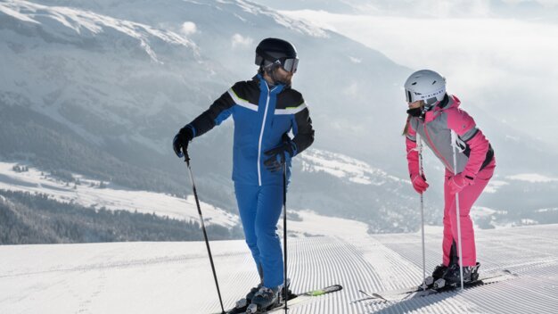 Zwei Skifahrer auf einer Skipiste vor schneebedeckten Bergen.
