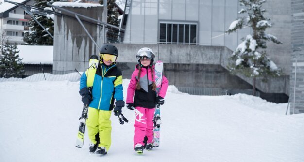 Two children in ski attire carrying skis in the snow. | © McKINLEY