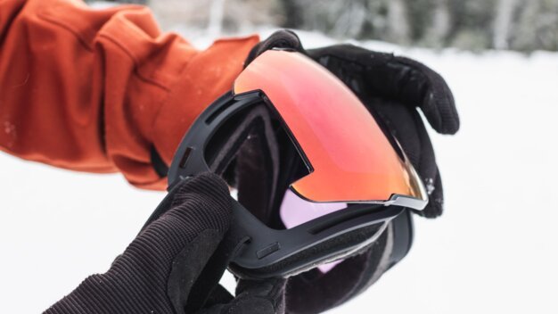 A person holds ski goggles with an orange lens in gloved hands.