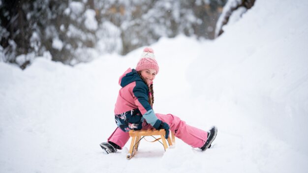 Mädchen sitzt auf einem Holzschlitten im Schnee und lächelt | © McKINLEY