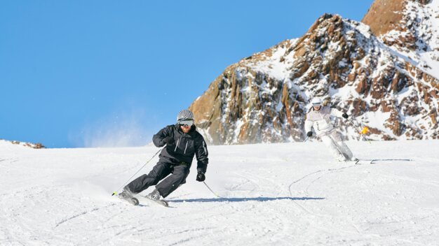 Zwei Skifahrer auf einer schneebedeckten Piste vor einer Bergkulisse.