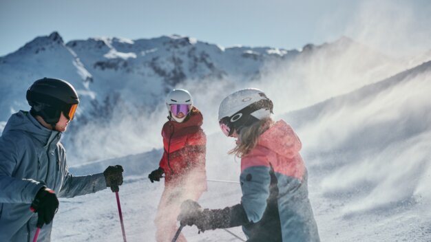 Three skiers wearing helmets and winter jackets stand on a snowy mountain slope, powder snow swirling around them, sunny mountain peaks in the background. | © INTERSPORT International Corporation GmbH