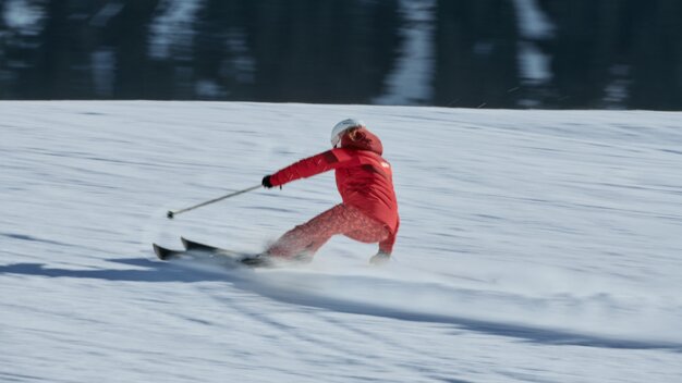 Person in roter Skijacke und hellem Helm fährt eine schnelle Kurve auf verschneiter Piste, Schnee spritzt, dunkler Wald im Hintergrund. | © INTERSPORT International Corporation GmbH