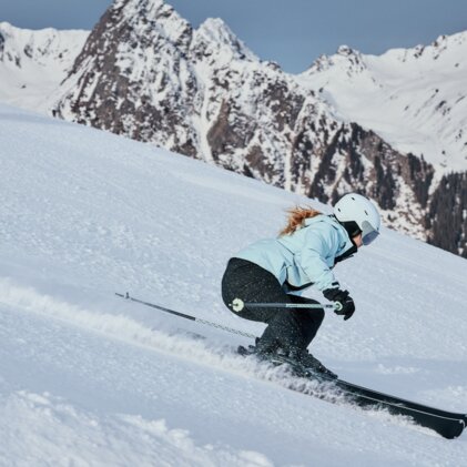 Frau beim Skifahren in hellblauer Jacke und weißem Helm, sie fährt eine steile, verschneite Piste vor schneebedeckten Bergen. | © INTERSPORT International Corporation GmbH