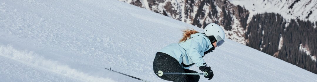 Femme en train de skier, vêtue d'une veste bleu clair et d'un casque blanc, elle descend une piste enneigée escarpée devant des montagnes enneigées. | © INTERSPORT International Corporation GmbH