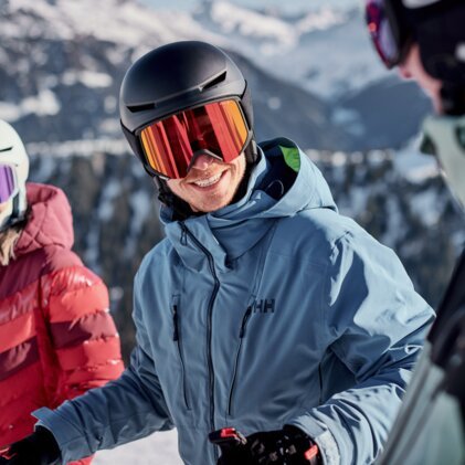 Three skiers wearing helmets and ski goggles on a snowy mountain slope, with a smiling man in a blue jacket in the foreground.