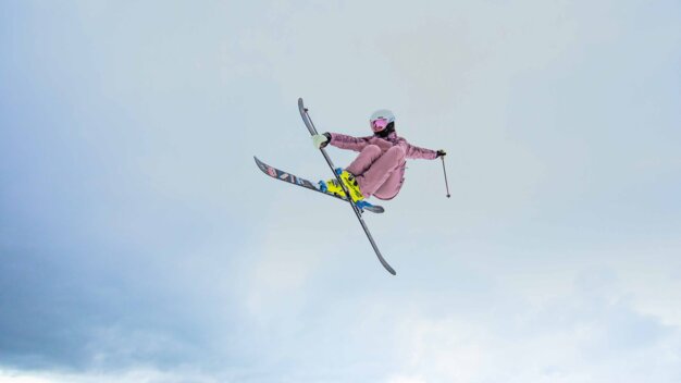 Skifahrende in rosa Skianzug mit Helm und rosa Skibrille führt bei einem Luftsprung gekreuzte Skier vor vor hellem Winterhimmel. | © INTERSPORT International Corporation GmbH