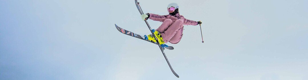 Skiers in pink ski suits with helmets and pink ski goggles perform a jump with their skis crossed against a bright winter sky. | © INTERSPORT International Corporation GmbH