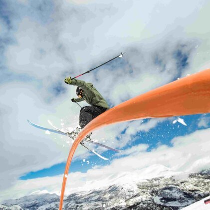 Skieur vêtu d'une veste verte et d'un casque sautant par-dessus un tube orange courbé dans un snowpark ; neige, montagnes et ciel nuageux en arrière-plan. | © INTERSPORT International Corporation GmbH