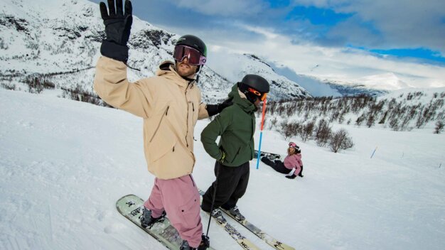 Three winter sports enthusiasts on a snow-covered slope in front of mountains: in the foreground, a snowboarder wearing a beige jacket and pink pants waves, next to him a skier in a green jacket stands, and in the background a skier in a pink jacket sits; all wearing helmets and ski goggles. | © INTERSPORT International Corporation GmbH