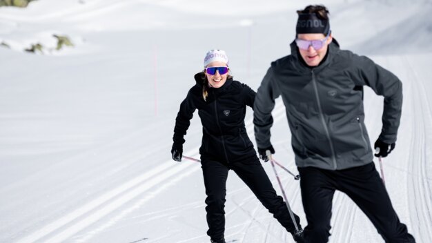 Two cross-country skiers in black outdoor clothing and protective goggles skiing one behind the other on a groomed trail in the sunshine, woman smiling. | © INTERSPORT International Corporation GmbH