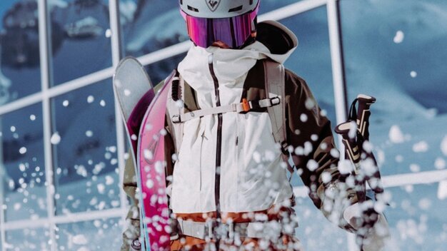 Skifahrende in weißer Jacke, Helm und violetter Skibrille trägt rosa Skier und Stöcke; aufspritzender Schnee vor Glasfassade mit Bergreflexion. | © INTERSPORT International Corporation GmbH