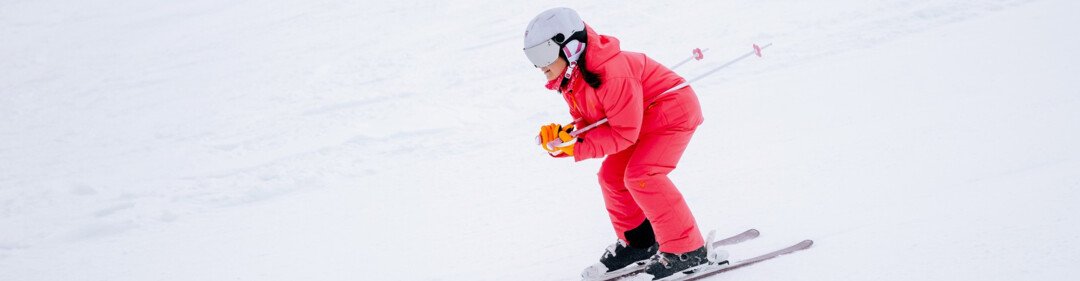 Person in rotem Skianzug und weißem Helm in leicht geduckter Haltung beim Skifahren auf verschneiter Piste, trägt orange Handschuhe und Skistöcke. | © INTERSPORT International Corporation GmbH