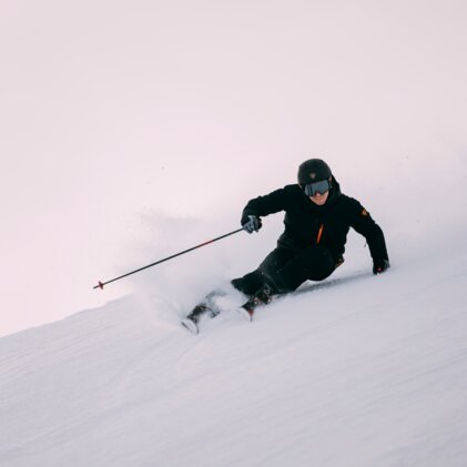 Person carving in deep snow, wearing a black helmet, ski goggles, black jacket, and skis. | © INTERSPORT International Corporation GmbH