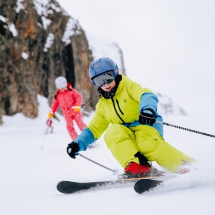 Child skiing: neon yellow ski suit, blue helmet, and red ski boots cutting turns on the slope, second person in red suit in the background in front of a rock face. | © INTERSPORT International Corporation GmbH