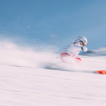 Fast-moving skier in a white jacket and red pants with orange skis, dynamic spray of snow against a blue sky. | © INTERSPORT International Corporation GmbH