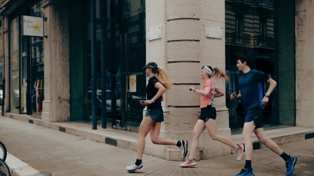 Three runners in sportswear (one in black, one in pink, one man in blue) run along the corner of a building on an urban shopping street. | © INTERSPORT International Corporation GmbH