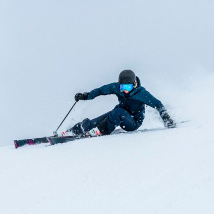 Skifahrer in dunkelblauer Jacke und schwarzem Helm mit blauen Skibrille, der in Pulverschnee eine tiefe Kurve fährt und Schnee aufwirbelt. | © INTERSPORT International Corporation GmbH