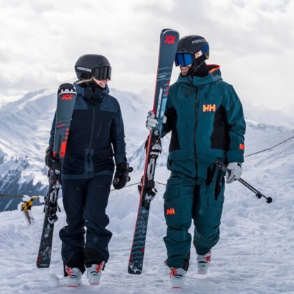 Zwei Skifahrer in Helmen und Skibrillen gehen nebeneinander durch tiefen Schnee, tragen jeweils Skier und Winterbekleidung (sichtbares HH‑Logo); verschneite Berglandschaft im Hintergrund. | © INTERSPORT International Corporation GmbH