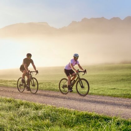 Zwei Radfahrer auf einer Schotterstraße, unterwegs durch eine neblige Landschaft im sanften Morgenlicht.