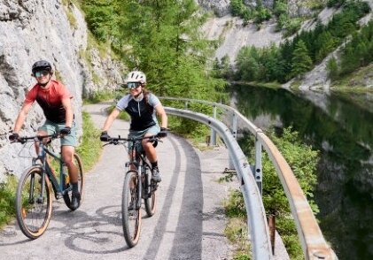 Zwei Radfahrer auf einem schmalen Weg entlang eines Flusses, eingebettet zwischen Felsen und dichter Natur.