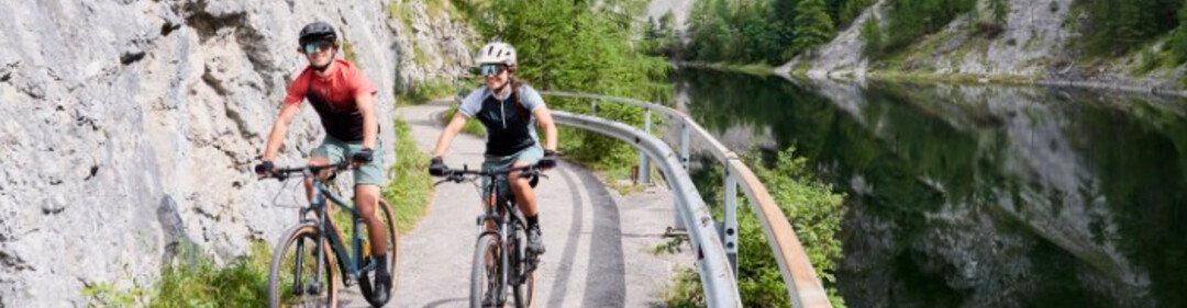 Zwei Radfahrer auf einem schmalen Weg entlang eines Flusses, eingebettet zwischen Felsen und dichter Natur.