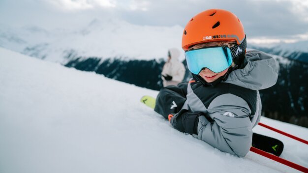 Child in orange ski helmet and blue goggles sitting in snow.