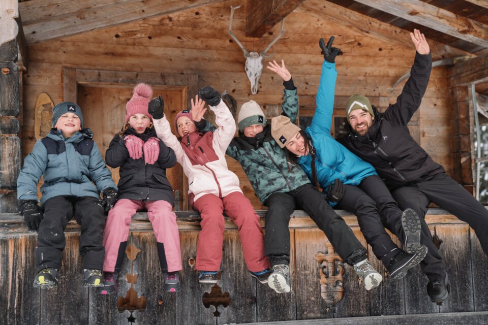 Gruppe von Personen in Winterkleidung sitzt vor einer Holzhütte und winkt. | © INTERSPORT International Corporation GmbH