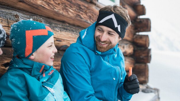 Père et fils se sourient en vêtements d'hiver devant une cabane en bois. | © IIC/McKINLEY