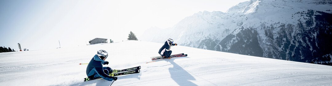 Zwei Skifahrer auf einer verschneiten Piste mit Bergen im Hintergrund. | © Fischer Sports GmbH