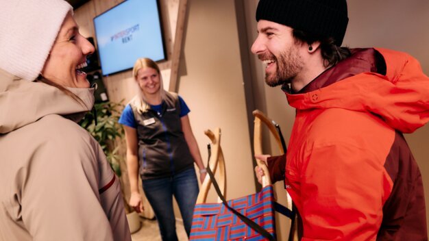 Laughing people with a sled at a ski rental shop. | © ARMIN WALCHER