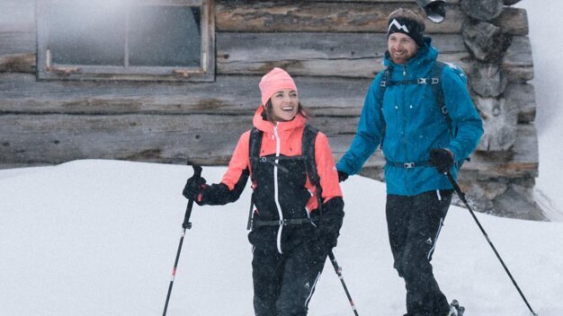 A couple snowshoeing in front of a wooden house. | © IIC/McKINLEY