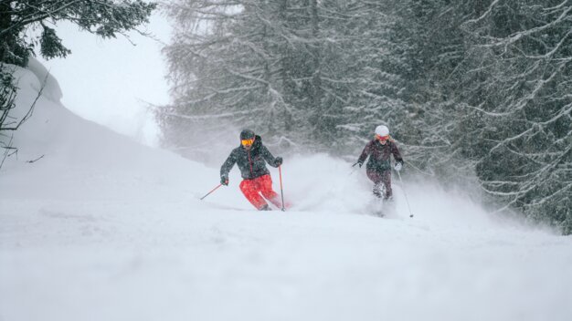 Two skiers skiing down a snowy slope in falling snow.