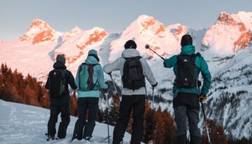 Vier Skifahrer betrachten eine schneebedeckte Bergkette bei Sonnenaufgang. | © Clément Hudry