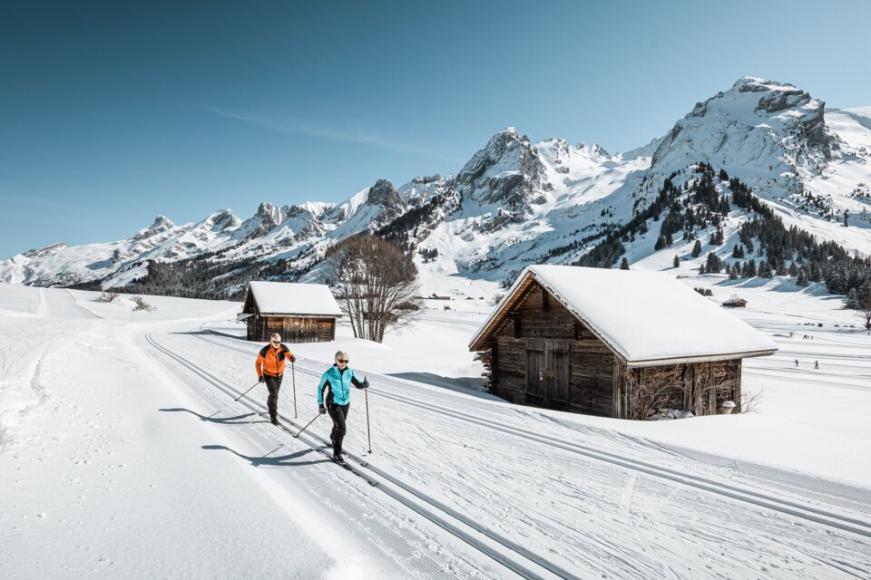 Two cross-country skiers on a trail in a snowy landscape with wooden huts and mountains in the background. | © C.HUDRY