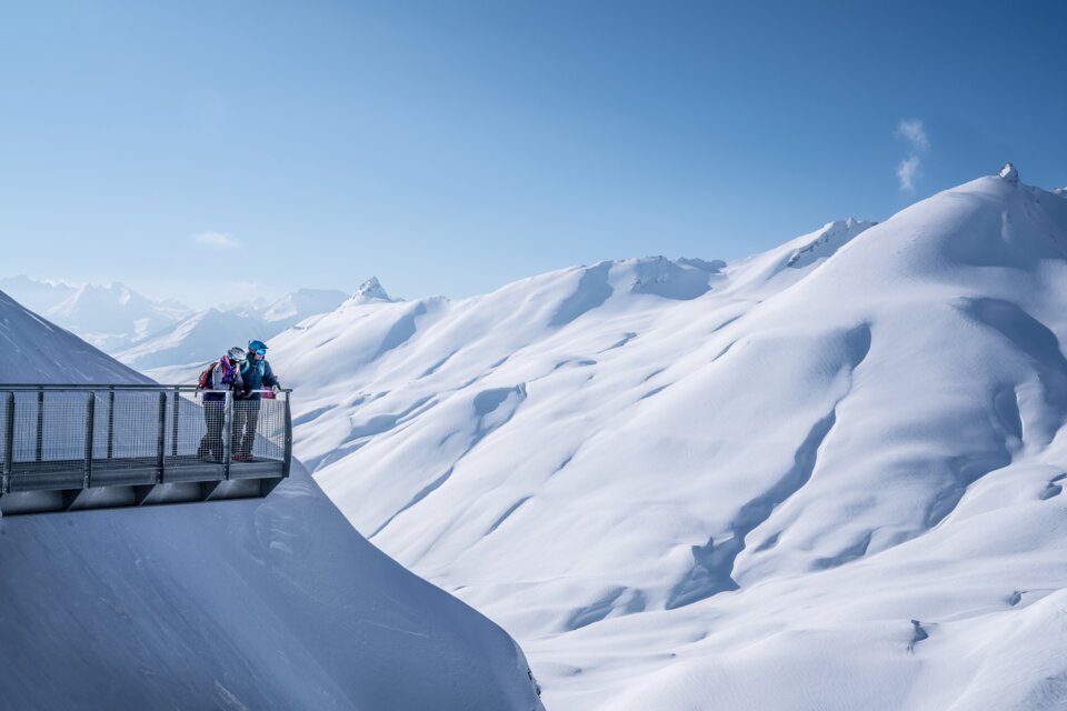 Zwei Personen auf Aussichtsplattform in verschneiter Berglandschaft.