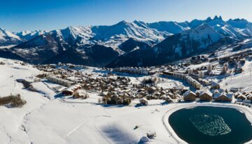 Panorama eines verschneiten Bergdorfes mit See und schneebedeckten Bergen im Hintergrund | © La Toussuire / A. Pernet