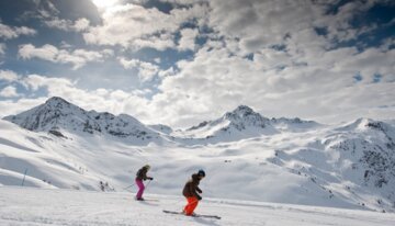 Two skiers on a slope with snowy mountains in the background. | © Gilles Lansard