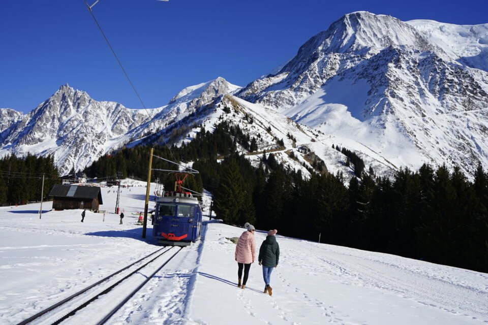Zwei Frauen gehen neben einer blauen Zahnradbahn in einer verschneiten Berglandschaft. | © OT Vallée de Chamonix / MR
