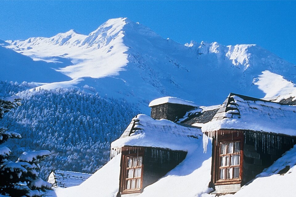 Snow-covered mountain lodge with snow-capped mountains in the background. | © OT Saint Lary