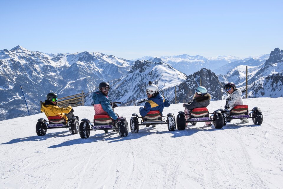 Group of people on Mountain Karts in the snow with mountains in the background | © Serre Chevalier Vallée Briançon - @laurapeythieu