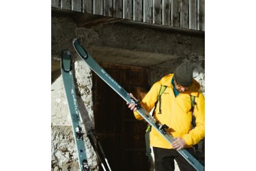 Man in yellow jacket holding skis in front of a wooden door.