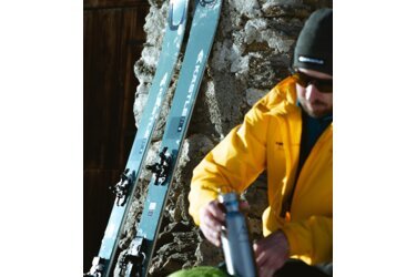 Skis and man with water bottle against a stone wall.